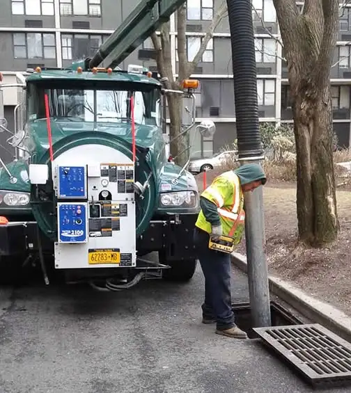 Green A&L vacuum truck with boom hose lowered into a catch basin, a worker in hi-vis guiding it in